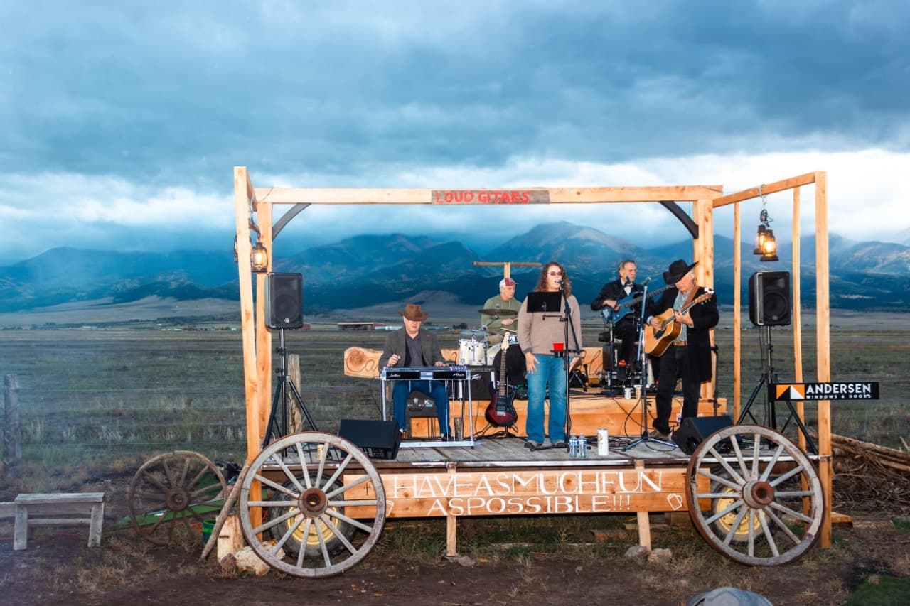 The full 5-piece band playing live on the completed Cadillac Wanch stage at dusk, with the Sangre de Cristo mountains and Andersen Windows & Doors signage in the background