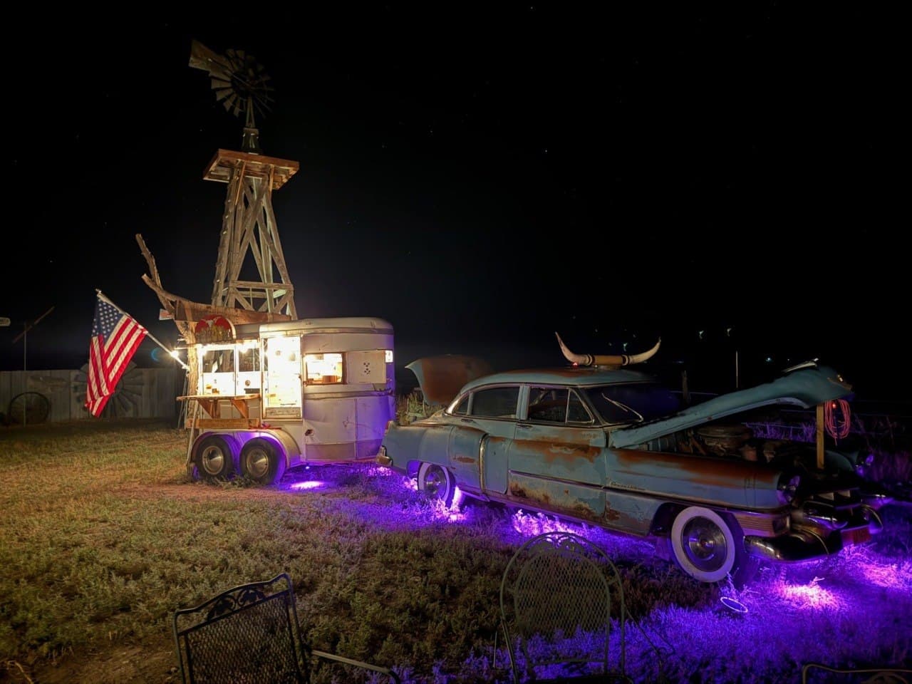 The Cadillac Wanch at night — vintage Cadillac with longhorn horns, saloon trailer, windmill, and purple lighting under a star-filled Colorado sky