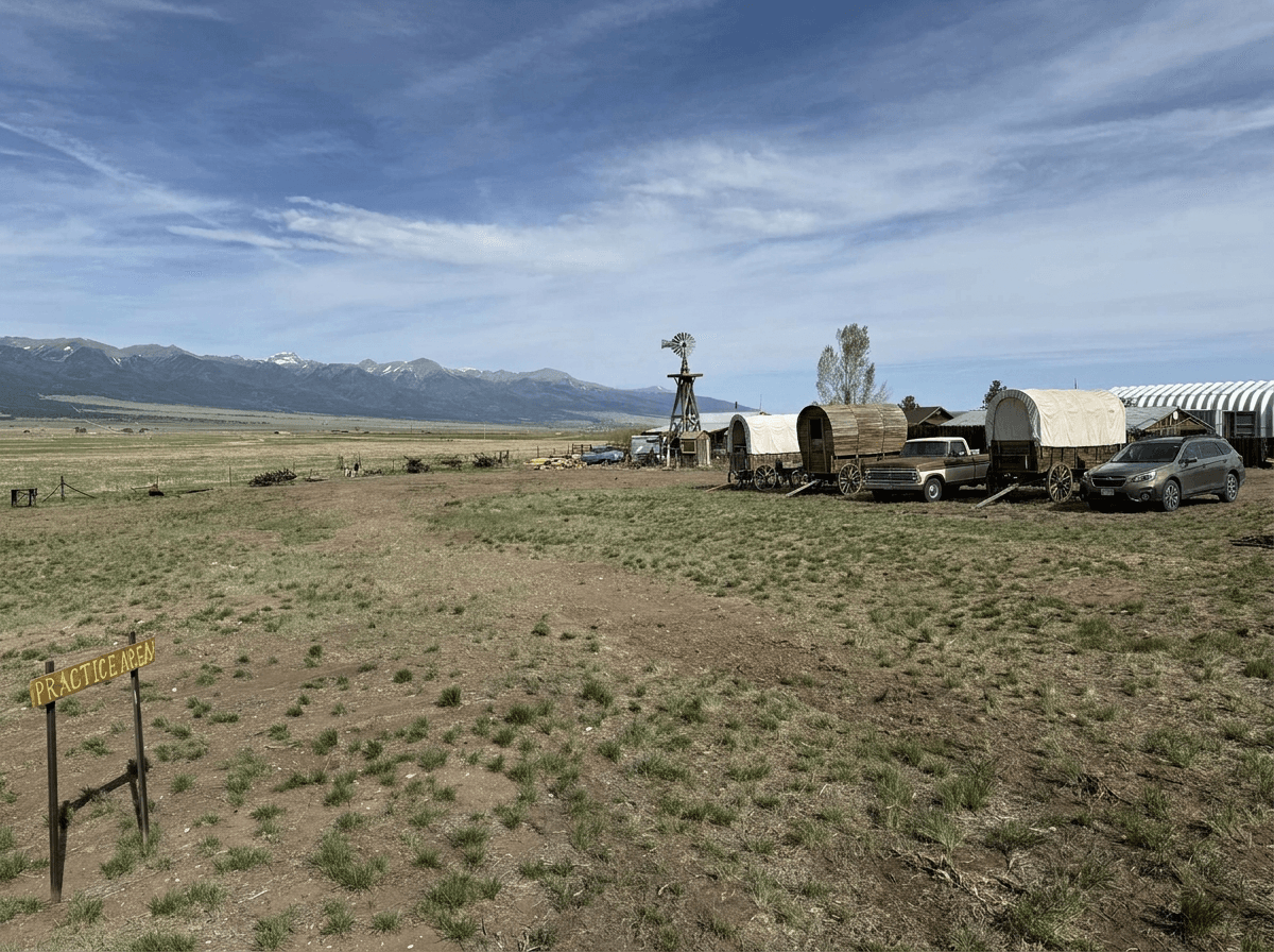 Existing practice area: open grassy field with a hand-painted 'PRACTICE AREA' sign, sheepherder wagons, a windmill, and a pickup truck in the distance, Sangre de Cristo mountains on the horizon