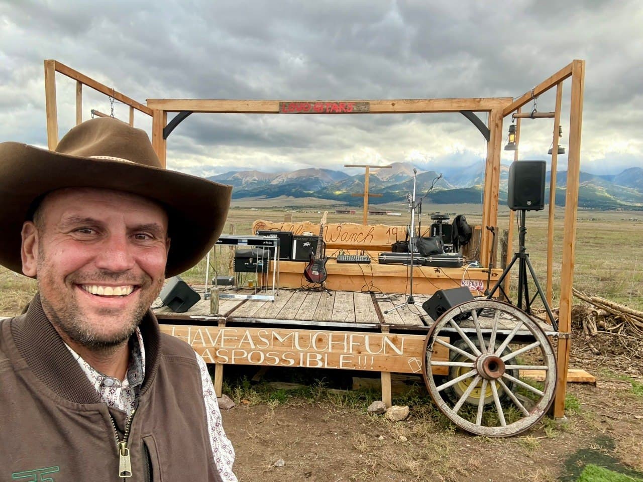 Devon in front of the Loud Guitars stage set up for a full five-piece band, with amps, guitar, and PA speakers, framed by the Sangre de Cristo mountains under a fall sky