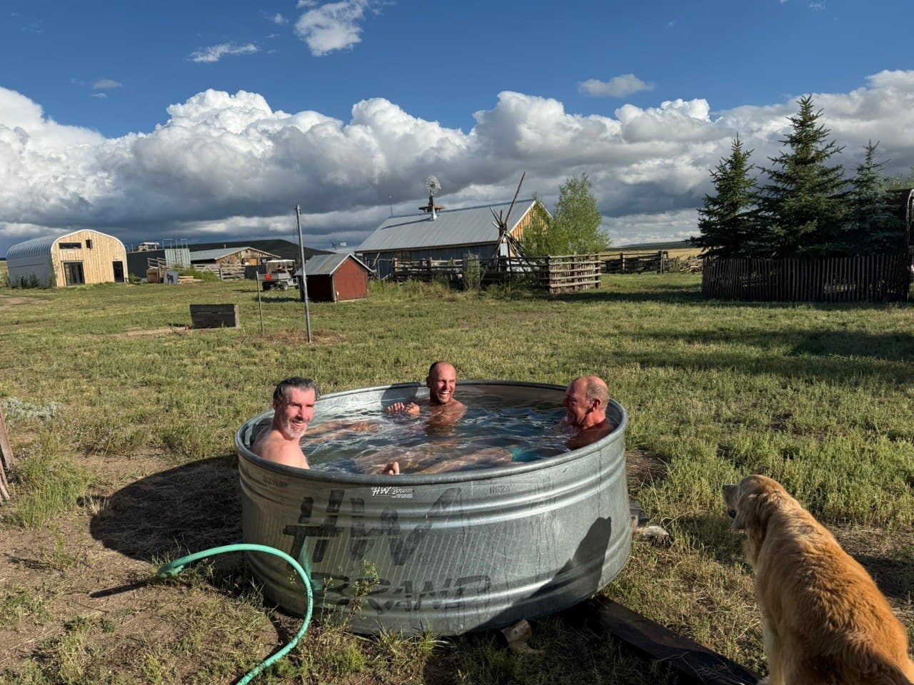 Wellness Center stock-tank cold plunge today — guests soaking with mountain pasture, barn, and windmill in the background