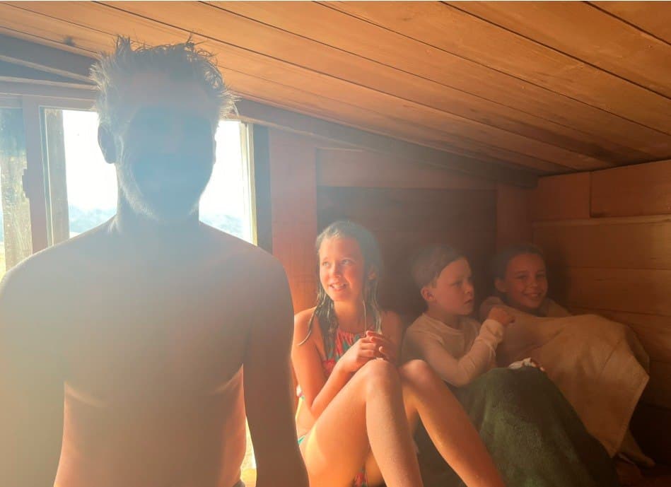 Family sauna session in daytime light — kids and dad warming up with mountain views through the window